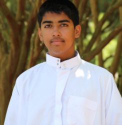 A young hafiz stands against a background of myrtle trees wearing a white thobe.
