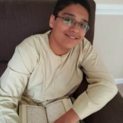 Young Hafiz sits in a chair, wearing a white kameez and holding Quran in his lap.