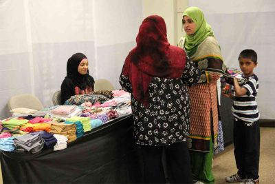 Vendor sits at booth during Eid Bazaar as women talk about scarf options.