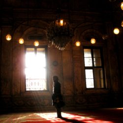 A person stands alone near the light of a window in a large, high-vaulted room with large, softly-lit lanterns suspended overhead.